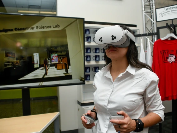 Woman wearing a white shirt uses a VR headset and handheld controllers. A large screen behind her displays a virtual scene labeled “Lundgren Consumer Science Lab.” Branded mugs and a red University of Arizona shirt are visible in the background.