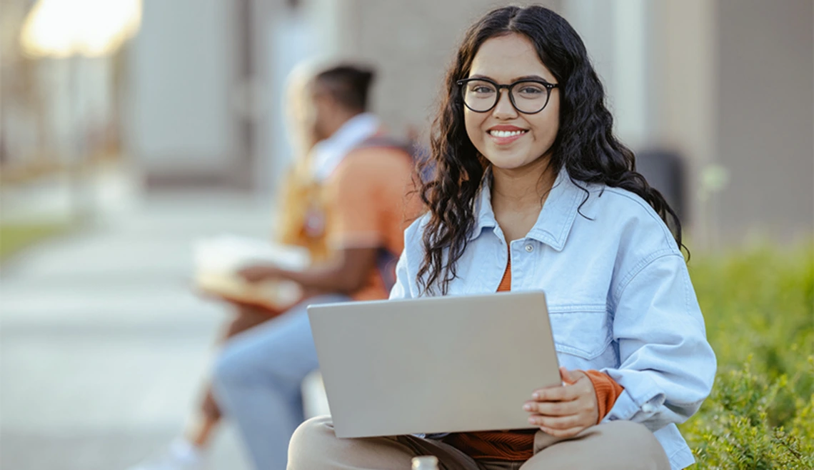 Smiling student holding laptop sitting outside