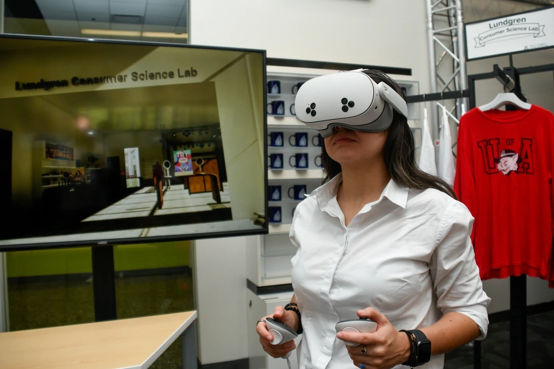 Woman wearing a white shirt uses a VR headset and handheld controllers. A large screen behind her displays a virtual scene labeled “Lundgren Consumer Science Lab.” Branded mugs and a red University of Arizona shirt are visible in the background.