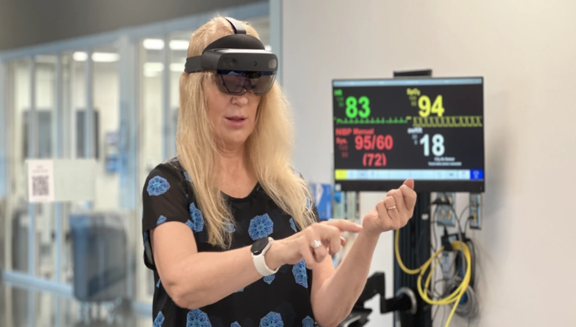 Janine Hinton wearing an augmented reality headset interacting with a virtual interface in a clinical simulation lab, with a medical monitor displaying vital signs in the background—demonstrating the use of immersive technology and AI in healthcare education.