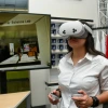 Woman wearing a white shirt uses a VR headset and handheld controllers. A large screen behind her displays a virtual scene labeled “Lundgren Consumer Science Lab.” Branded mugs and a red University of Arizona shirt are visible in the background.